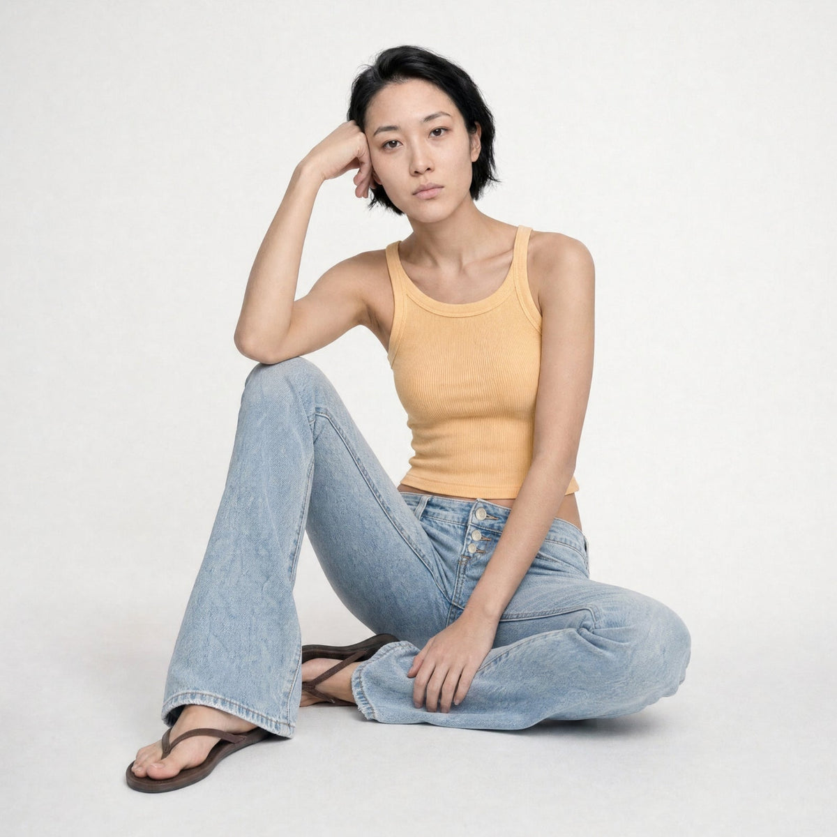 Rose model sitting on the floor wearing a yellow tank top and a light blue indigo Bubblegum Jeans on a white background