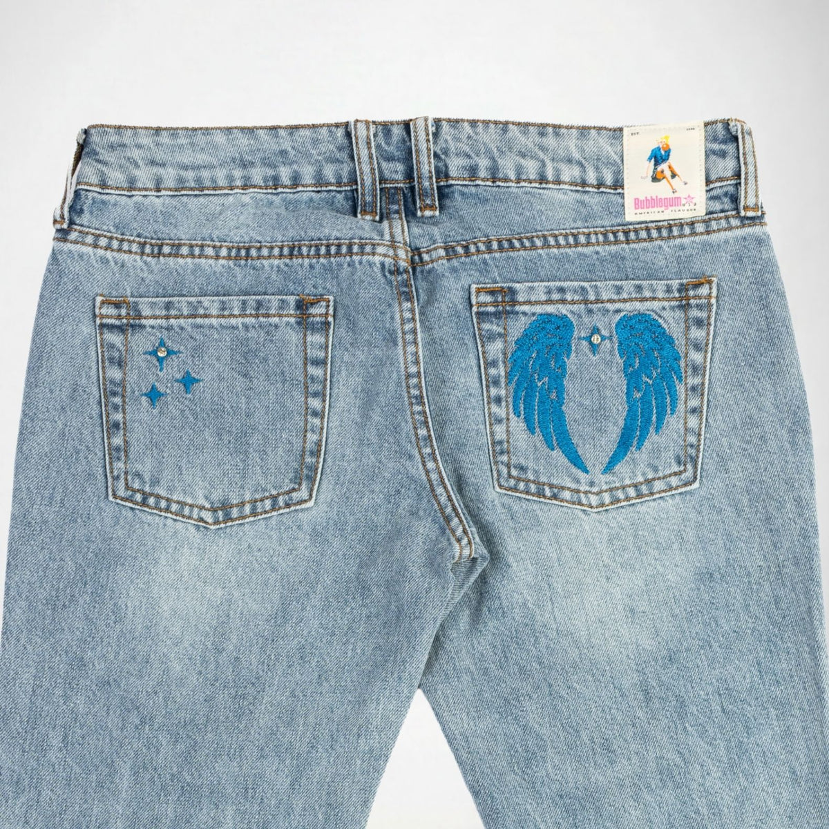Back view of light-wash blue denim Bubblegum jeans featuring embroidered blue designs on the back pockets, including wings and star embroidery on the right pocket and three shining stars embroidery on the left pocket, with a Bubblegum USA brand patch visible at the waistband.