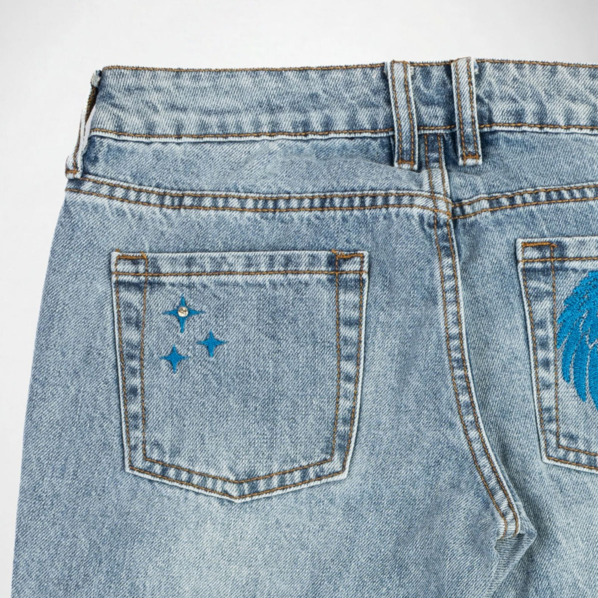 Close-up back view of light-wash blue denim Bubblegum jeans showing the waistband, belt loops, and back pockets with blue embroidered and rhinestone details.