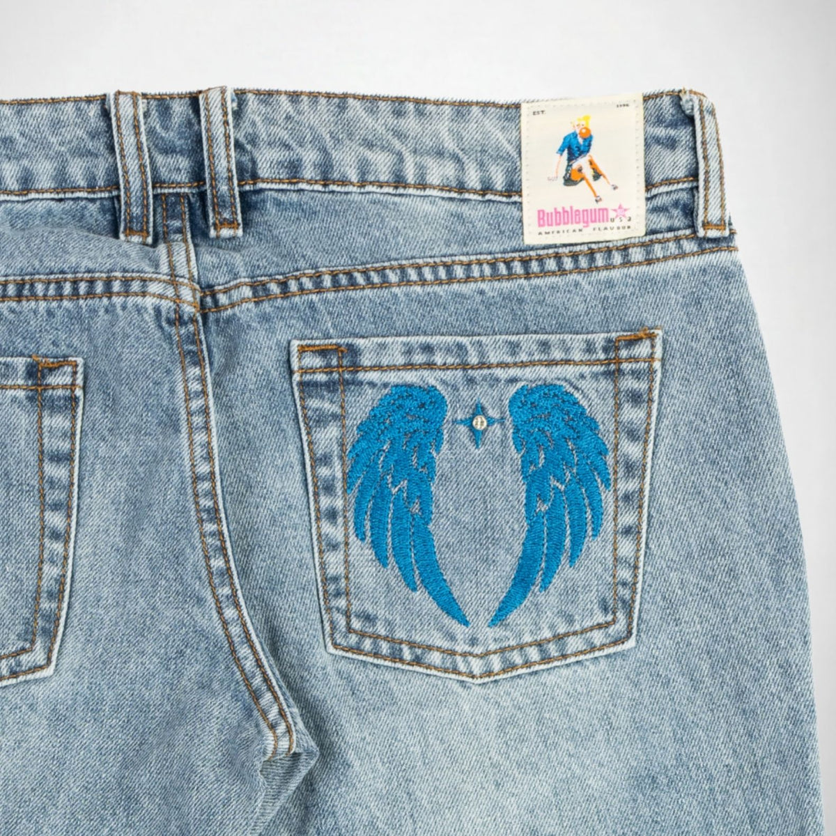 Close-up of the back of light-wash blue denim Bubblegum jeans showing a large embroidered pair of bright blue wings on the back pocket, with a visible Bubblegum brand label at the waistband.