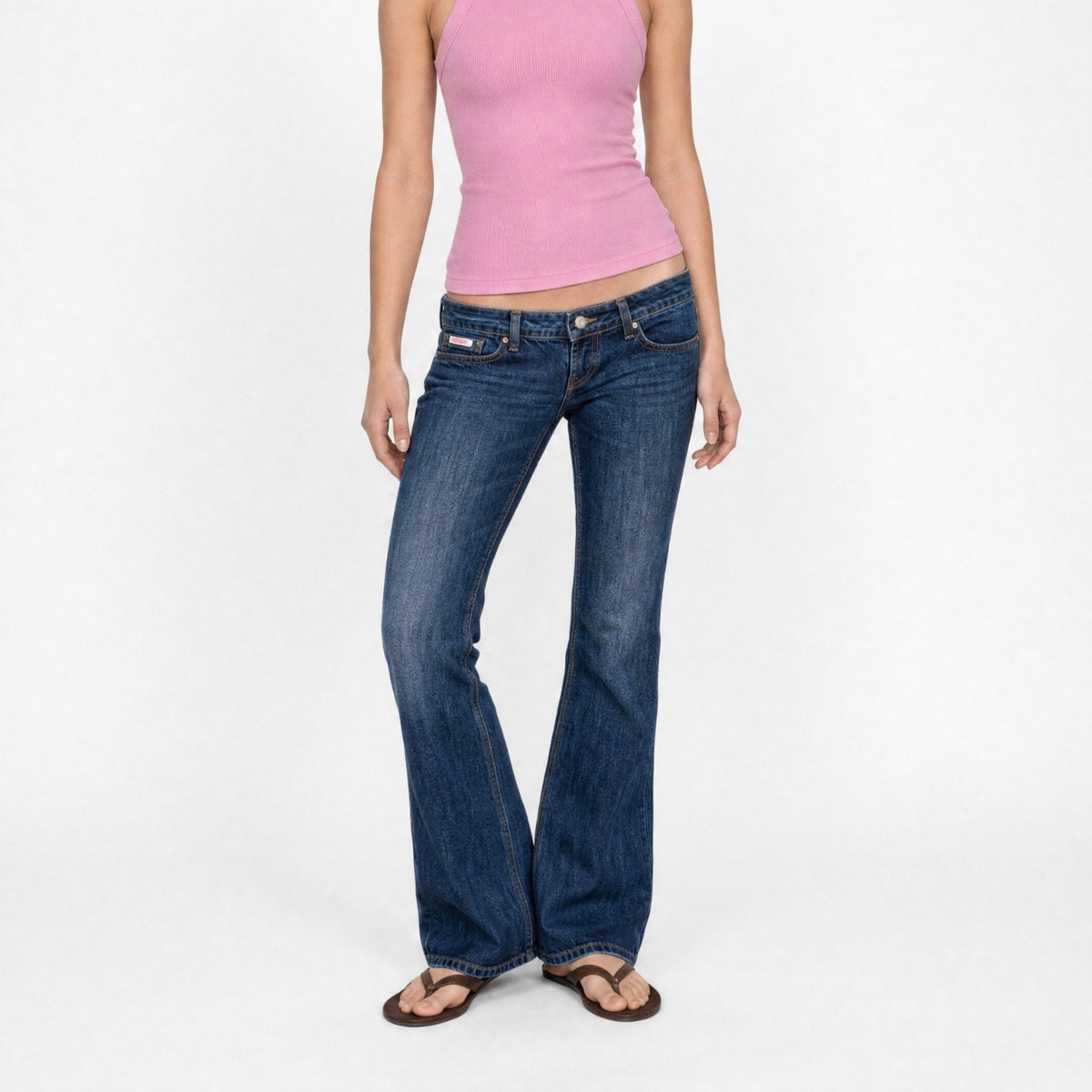 A model wearing a pink tank top and the Jane super low rise flare jeans in mid indigo vintage wash from Bubblegum USA brand, front of a white background.