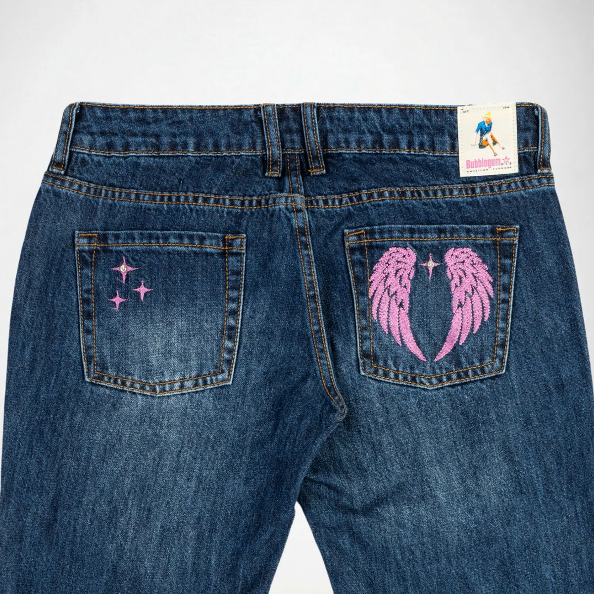 Back view of a pair of indigo denim, Bubblegum jeans featuring pink embroidered designs on the back pockets, including a pair of angel wings and shining  star on the right pocket, and three stars on the left pocket. A brand Bubblegum USA label is visible on the back waistband.