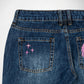 Close-up back view of dark blue denim Bubblegum jeans showing the back pockets with pink embroidered star and a shining rhinestone details on a white background.