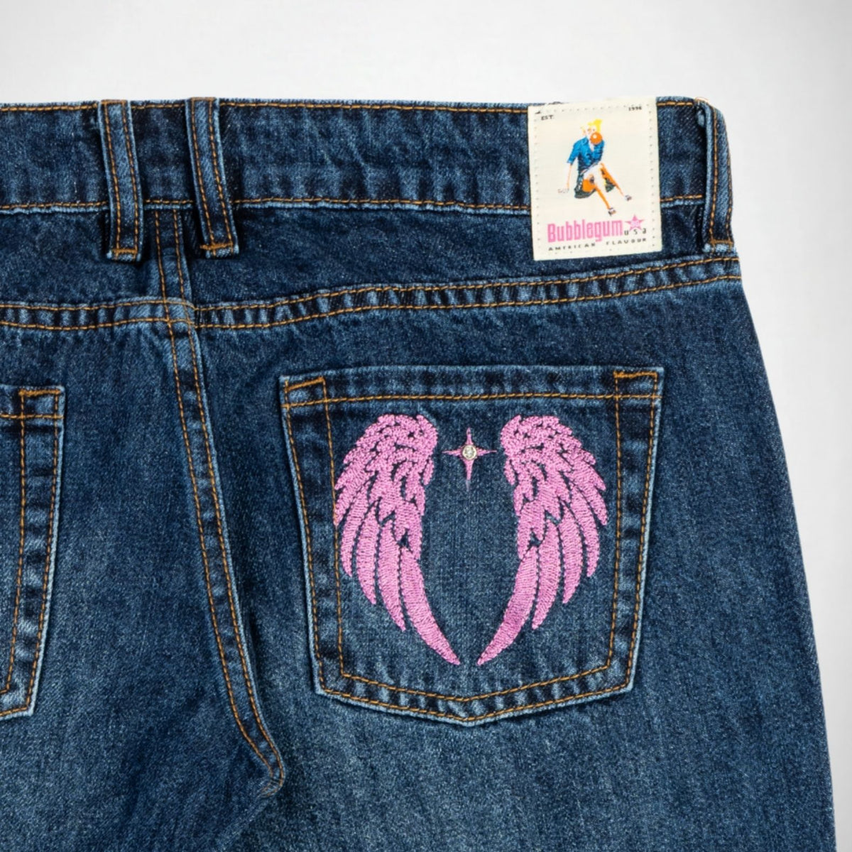 Close-up of the back of dark blue denim Bubblegum jeans showing a pink embroidered angel wings design on the back pocket, with a visible Bubblegum USA brand patch at the waistband.