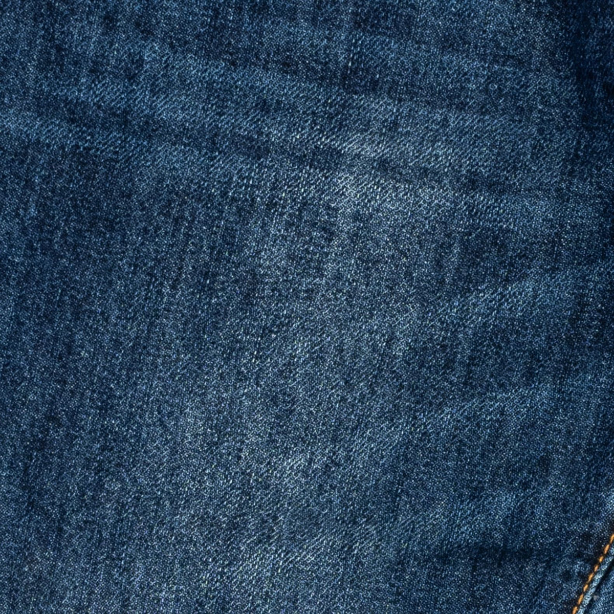 A close-up of dark blue denim fabric showing the detailed twill weave texture and a subtle yellow-orange contrast stitch in the bottom right corner.