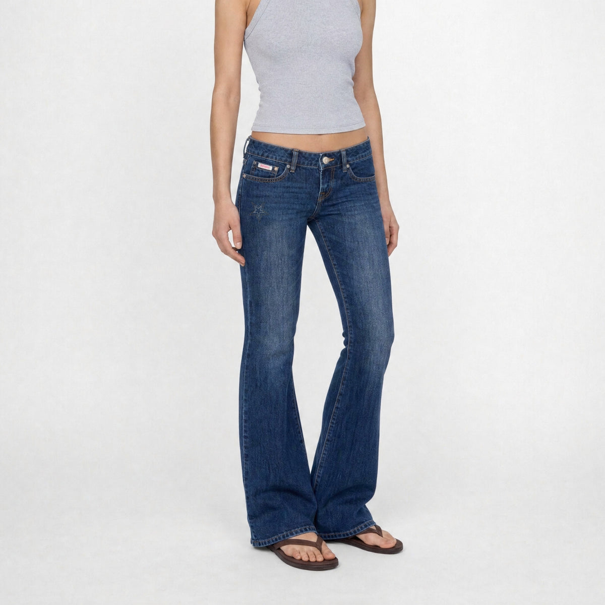 A model wearing a pink tank top and the Jane super low rise flare jeans in mid indigo vintage wash from Bubblegum USA brand, front of a white background.