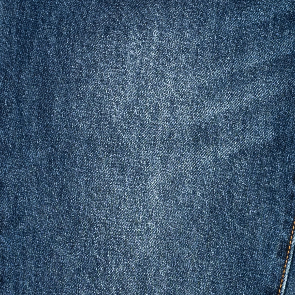 A close-up of blue denim fabric texture with a subtle diagonal twill weave and a small portion of orange contrast stitching visible at the bottom right.