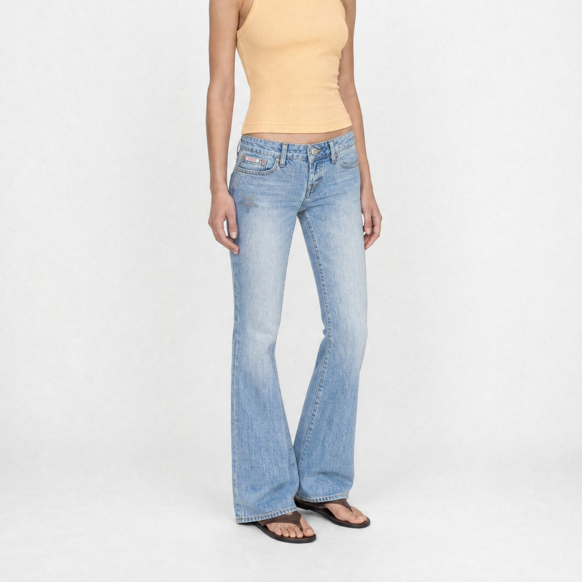 A model wearing Jane super low rise in light-wash blue Bubblegum jeans, a light orange ribbed tank top, and black sandals, standing against a plain white studio background.