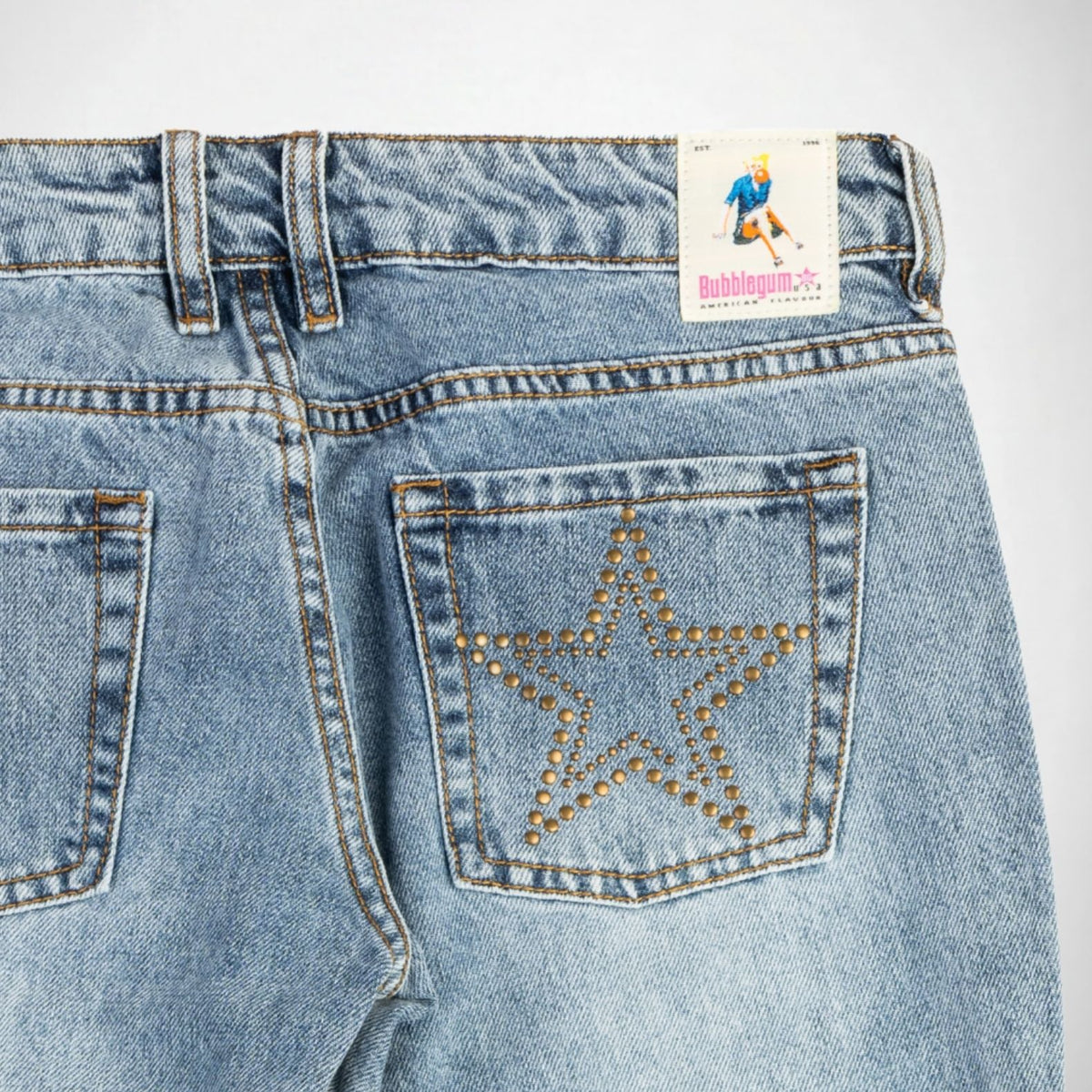 Close-up back view of light-wash blue denim Bubblegum jeans showing the waistband, belt loops, back pockets, and a large star-shaped stud embellishment outlined in gold on the right pocket. A Bubblegum USA brand patch is visible at the back waist.