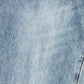 A close-up of light blue denim fabric with a subtle diagonal twill texture and a small portion of gold-toned stitching visible in the lower right corner.