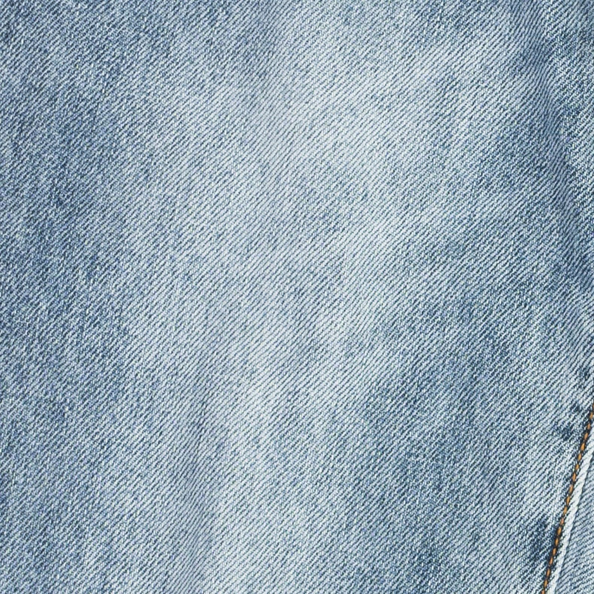 A close-up of light blue denim fabric with a subtle diagonal twill texture and a small portion of gold-toned stitching visible in the lower right corner.