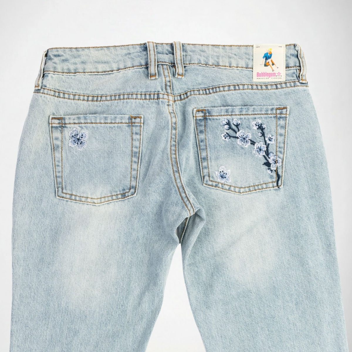 Back view of light-wash blue denim Bubblegum jeans with embroidered indigo and white cherry blossom flower details on both back pockets and a visible 'Bubblegum USA' logo patch at the waistband.