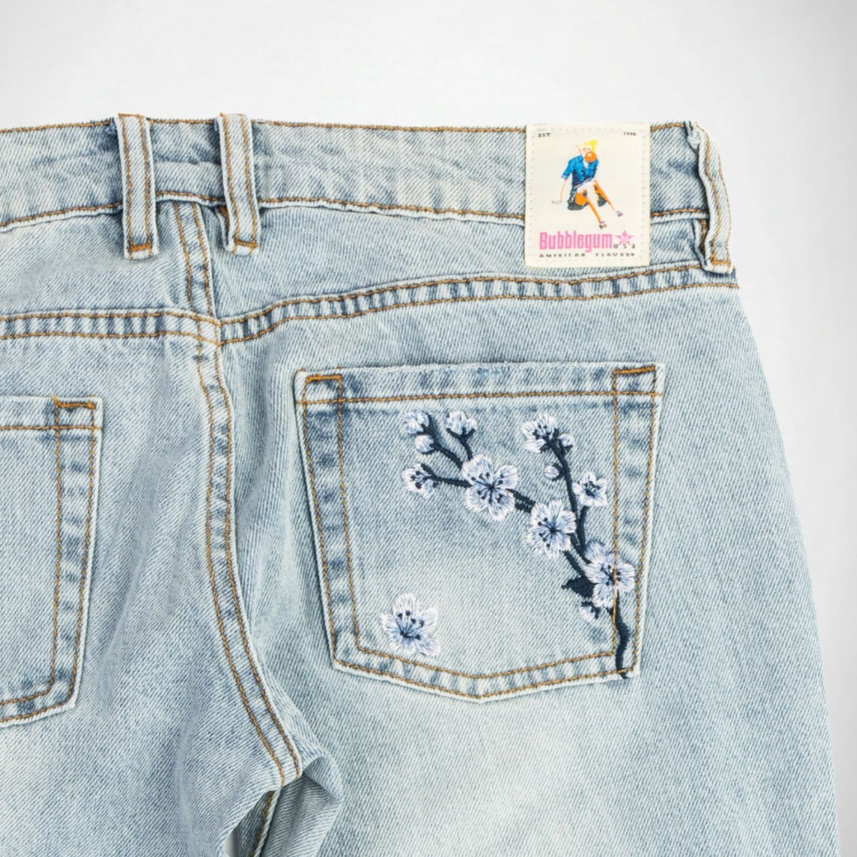 Close-up back view of light-wash denim Bubblegum jeans showing the back pocket with embroidered blue and white cherry blossom flower detail, plus a visible 'Bubblegum USA' label at the waistband.