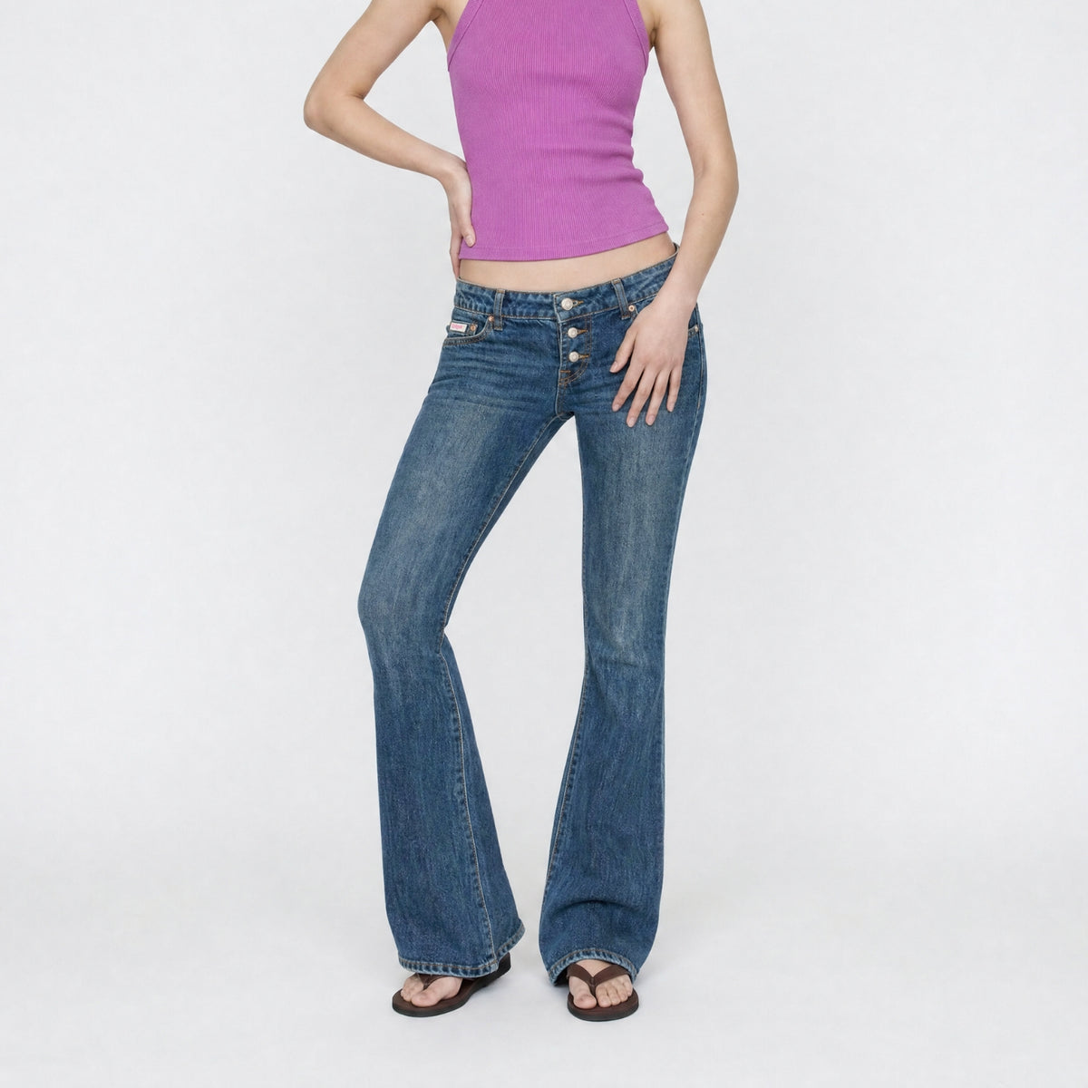 A model wearing the Rose super low rise, flare leg in indigo vintage wash Bubblegum jeans with a purple ribbed tank top and brown sandals, standing against a plain white studio background.