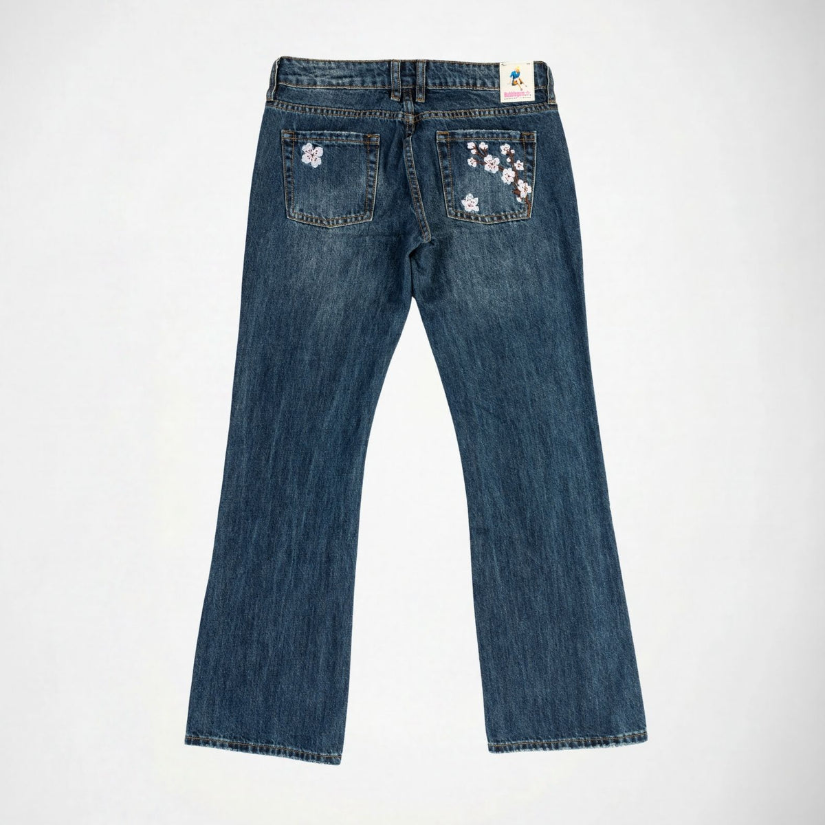 Back view of a pair of blue denim Bubblegum jeans with pink and white cherry blossom flower embroidery on the back pockets, displayed flat against a light gray background.