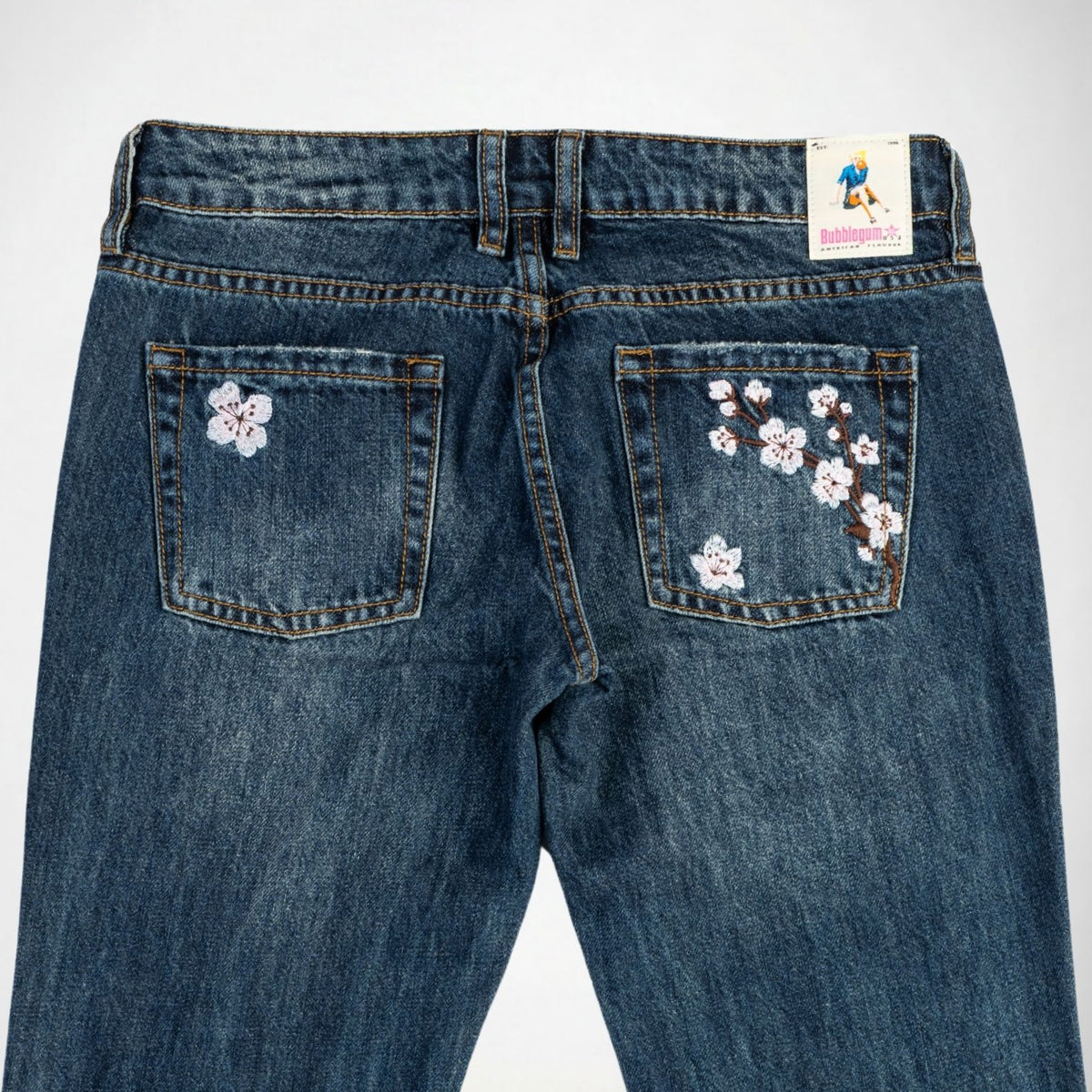 Back view of dark blue denim Bubblegum jeans featuring embroidered white and pink cherry blossom flowers on both back pockets, with a visible 'Bubblegum USA' brand label at the waistband.