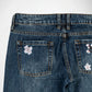 Close-up back view of dark blue denim Bubblegum jeans showing the waistband, belt loops, and back pockets with embroidered white and pink cherry blossom flowers on a light gray background.