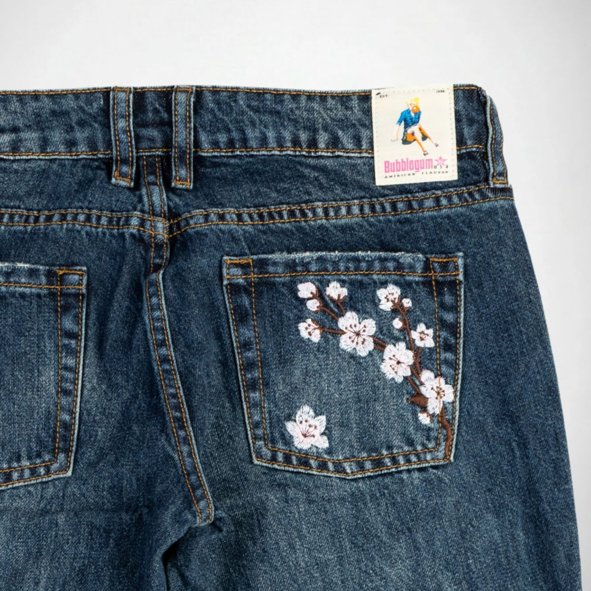 Close-up back view of dark blue denim Bubblegum jeans with embroidered Pink and white cherry blossoms on the right pocket. A sewn-on 'Bubblegum USA' brand patch is visible at the waistband.