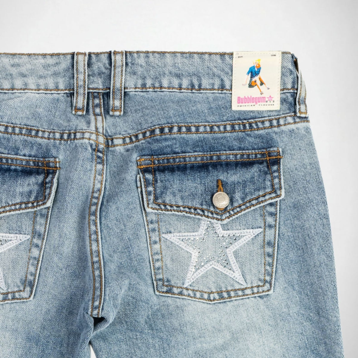 Close-up back view of light blue denim Bubblegum jeans showing the waistband, belt loops, rear pockets, a silver button with a white star embroidery with rhinestones embellishment, and a visible Bubblegum USA brand patch.