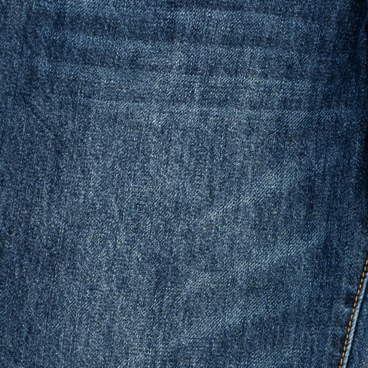 A close-up of dark blue denim fabric with a subtle diagonal twill texture and a small portion of orange contrast stitching visible on the right edge.