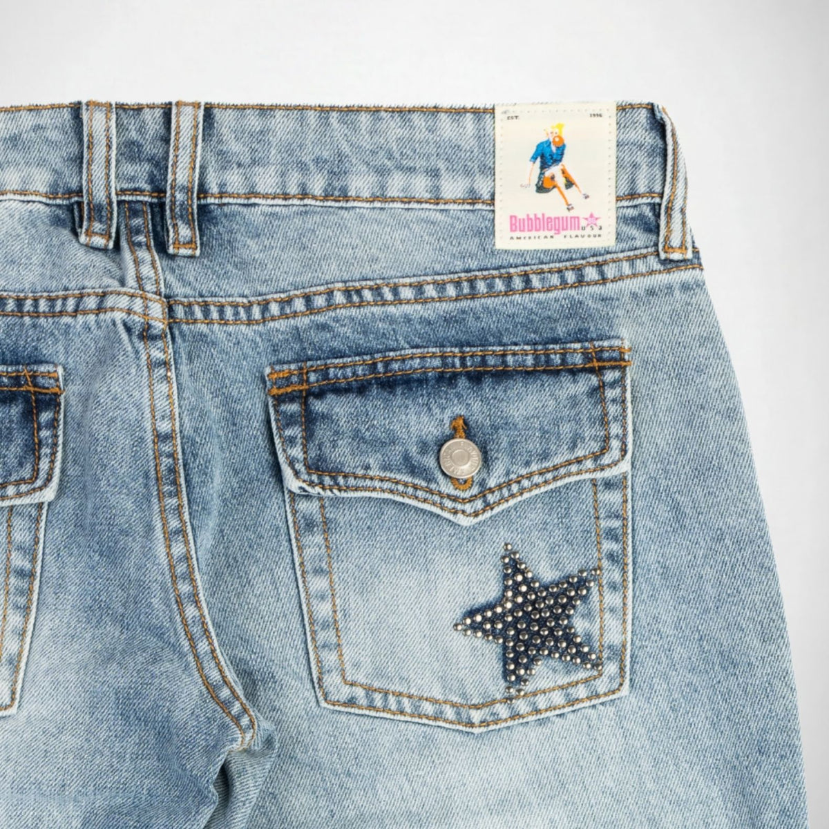 Close-up of the back waistband and pocket area of light-wash blue denim Bubblegum jeans. A square brand patch reading 'Bubblegum USA' is visible at the back waist, and a silver studs embellished star-shaped to the right back pocket.