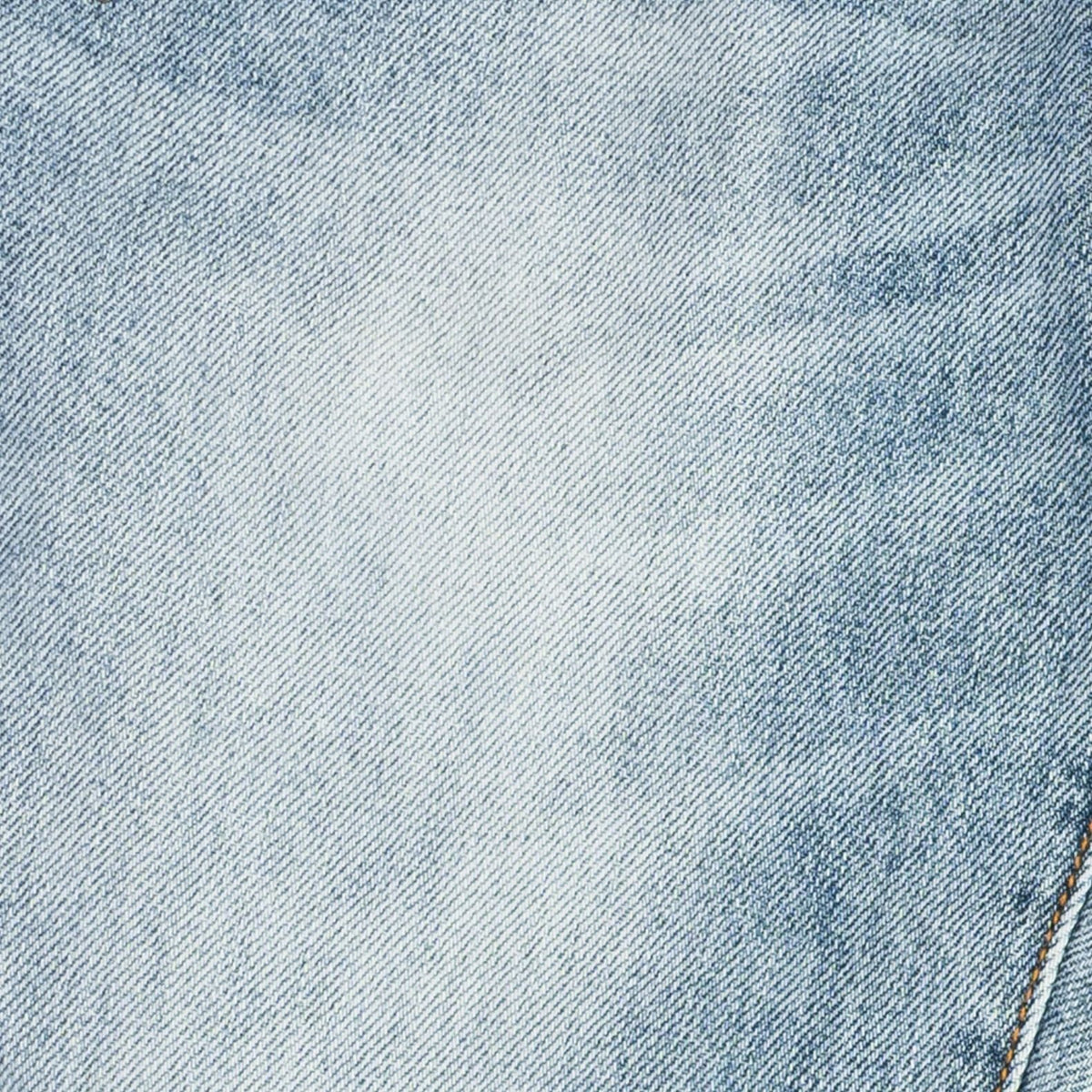 A close-up of light blue denim fabric with a subtle white faded area in the center and a small portion of tan stitching visible in the lower right corner.