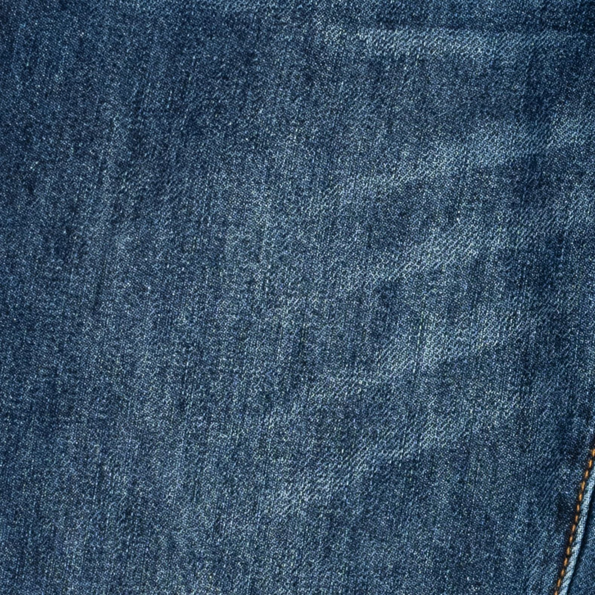 A close-up of dark blue denim fabric with a subtle diagonal twill texture and a small portion of orange-brown stitching visible in the lower right corner.
