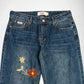 A pair of blue denim Bubblegum jeans featuring embroidered flowers on the left leg, photographed flat against a light gray background.
