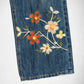 Close-up of a blue bubblegum denim garment, likely a skirt or pants leg, featuring embroidered floral details in orange, white, and yellow with green leaves on a white background.