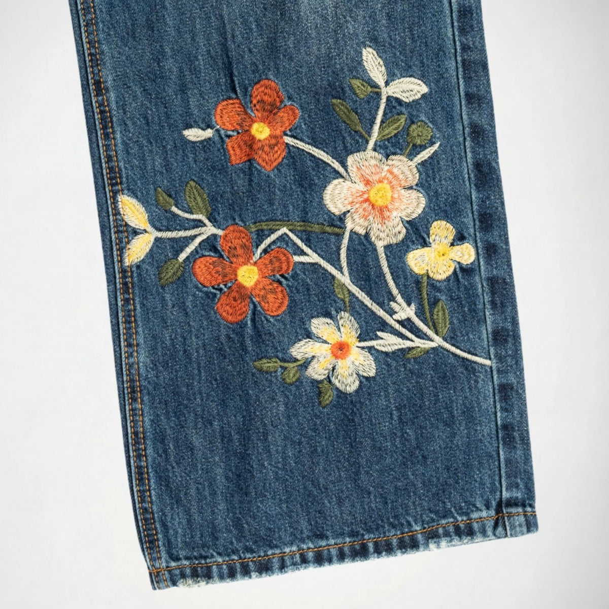 Close-up of a blue bubblegum denim garment, likely a skirt or pants leg, featuring embroidered floral details in orange, white, and yellow with green leaves on a white background.