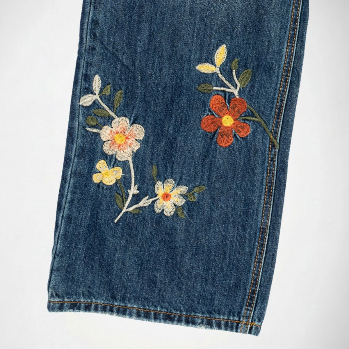 Close-up of a blue bubblegum denim garment leg with embroidered floral details in white, yellow, orange, and green on a plain light background.
