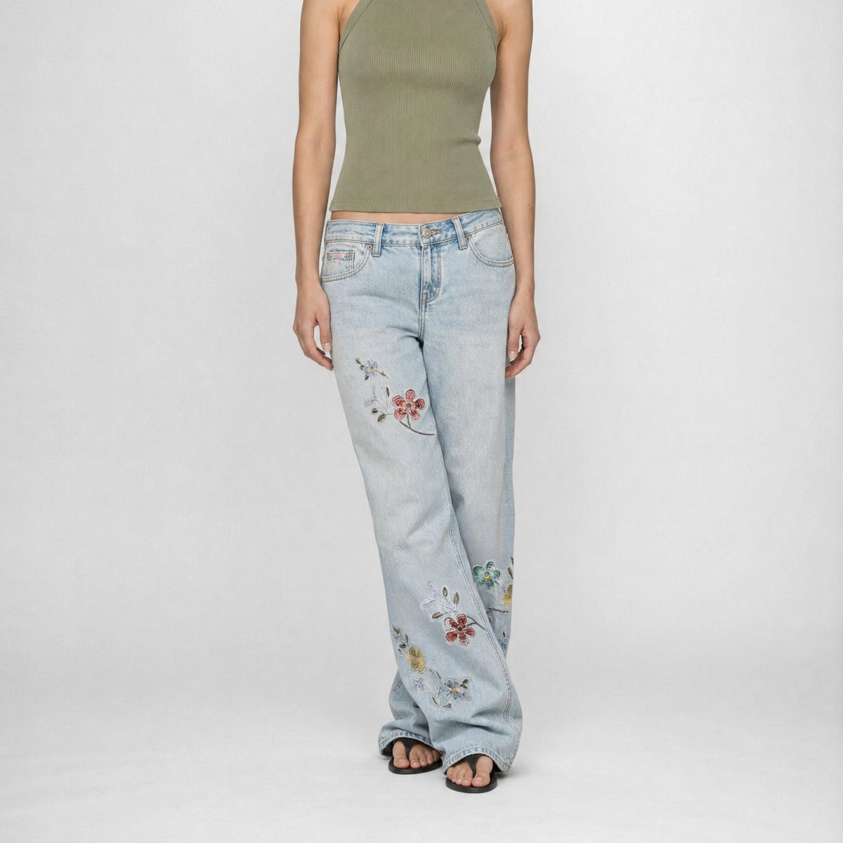 A model wearing the Lisa low baggy wide, mid rise in light-wash Bubblegum wide-leg denim jeans with colorful floral embroidery on the legs, paired with an olive green halter top and black sandals, against a plain white studio background.