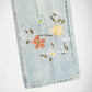 Close-up of a light blue Bubblegum denim garment with embroidered floral details in white, yellow, orange, and green on a plain white background.