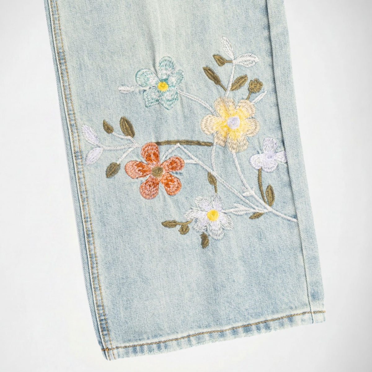 Close-up of a light blue Bubblegum denim garment with embroidered floral details in white, yellow, orange, and green on a plain white background.