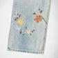 Close-up of a light blue Bubblegum denim garment, likely a pant leg, featuring embroidered floral details in yellow, orange, and white with green and brown leaves on a white background.