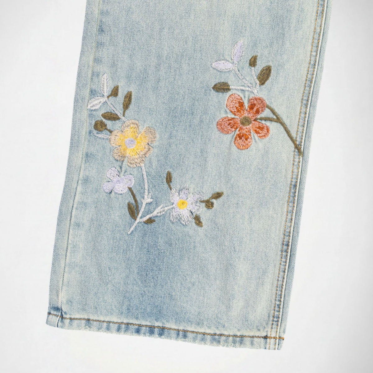 Close-up of a light blue Bubblegum denim garment, likely a pant leg, featuring embroidered floral details in yellow, orange, and white with green and brown leaves on a white background.