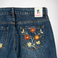 Close-up of the back waistband and right back pocket of blue denim Bubblegum jeans with colorful floral embroidery in orange, yellow, and white on the pocket. A Bubblegum USA brand label is visible at the back waistband.