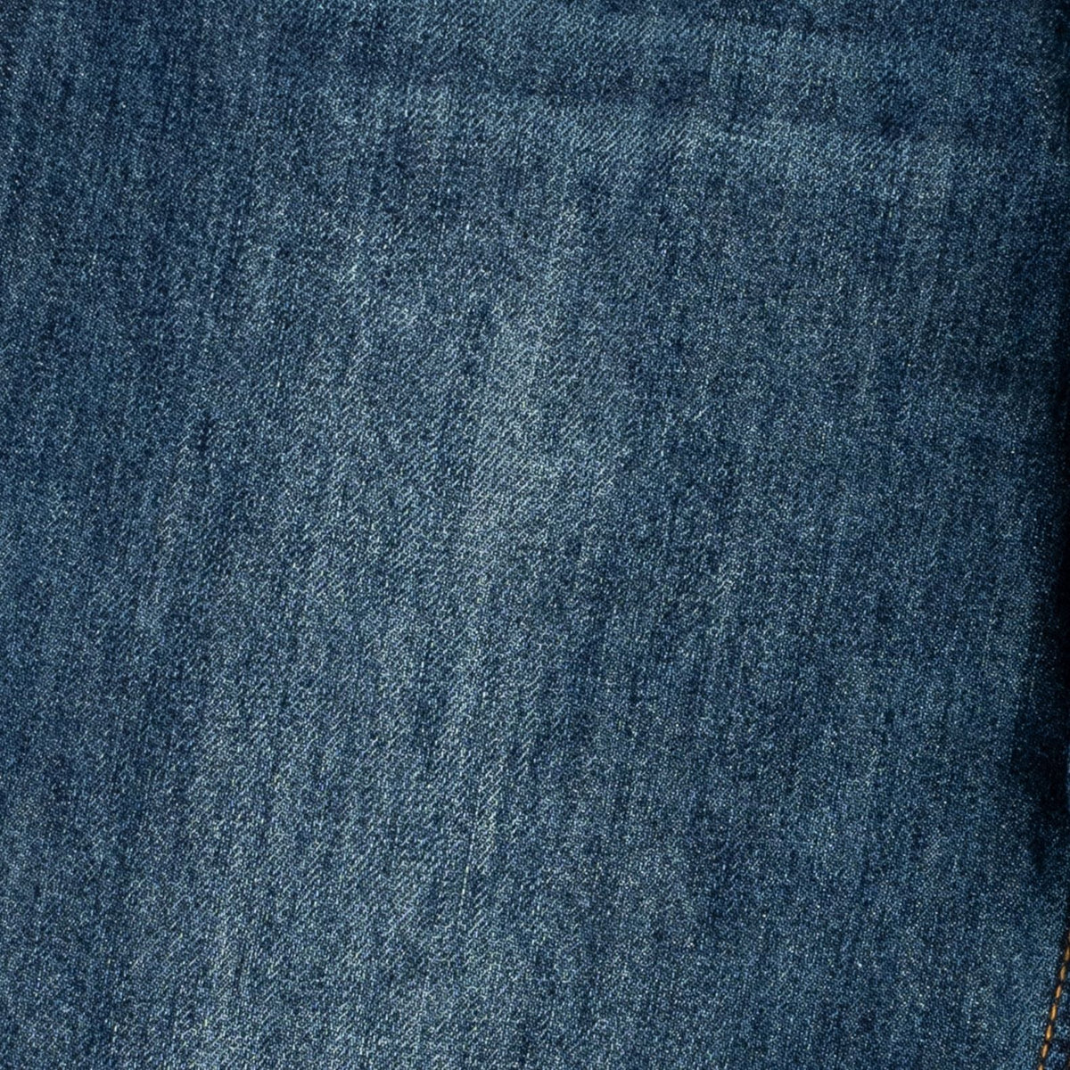 A close-up of dark blue denim fabric texture with subtle tonal variation and a small portion of contrasting stitching visible at the lower right edge.