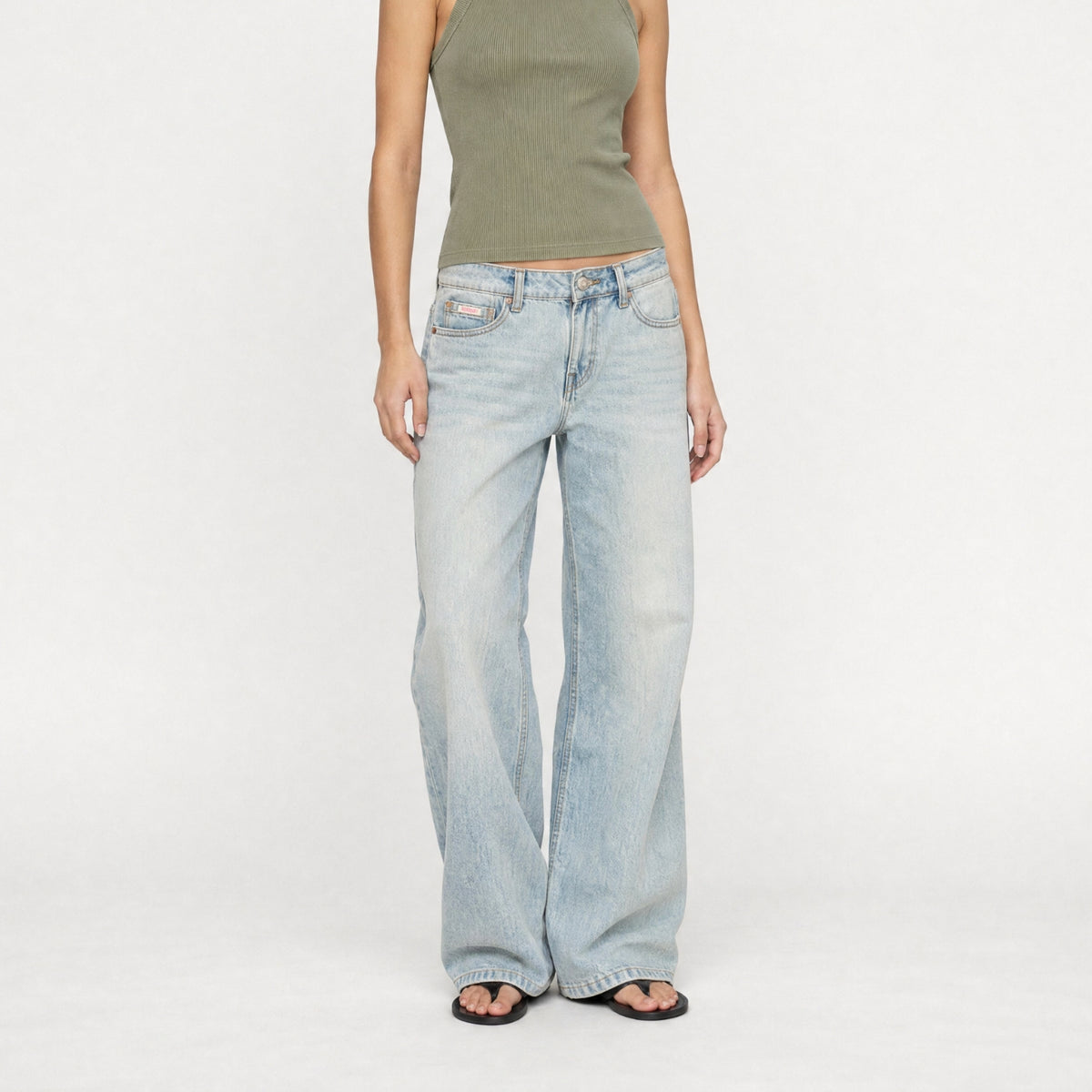 A model wearing the Lisa low baggy wide in mid-rise light-wash, wide-leg Bubblegum denim jeans with a ribbed olive green tank top and black sandals, standing against a plain white studio background.