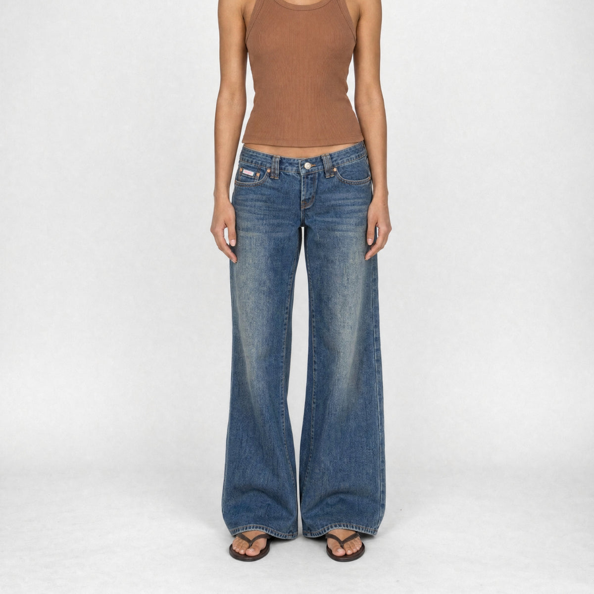 A model wearing the Luna super low wide in indigo dirty vintage wash, wide-leg Bubblegum jeans, a brown ribbed tank top, and brown sandals, standing against a light gray studio background.