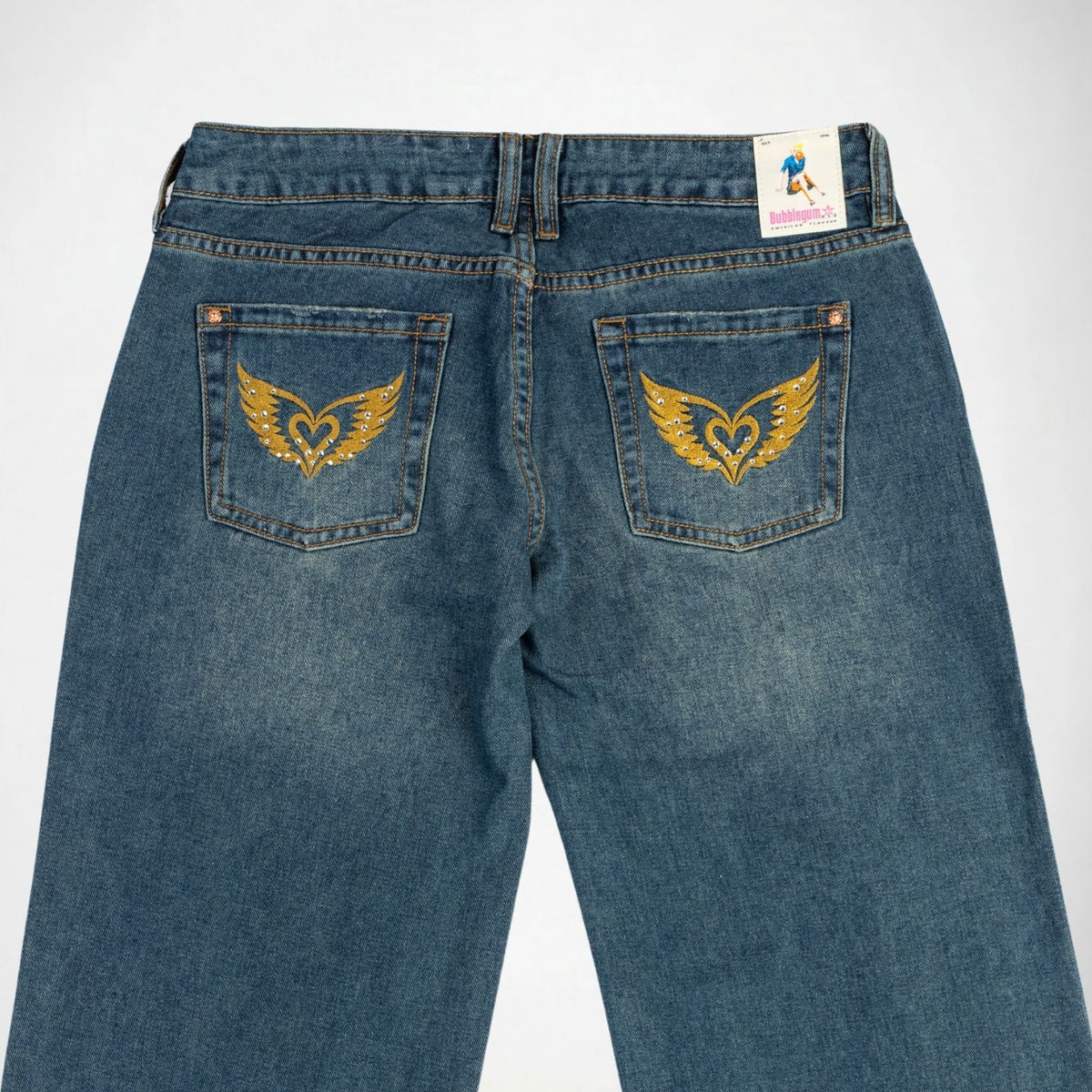 Back view of a pair of blue denim Bubblegum jeans featuring a winged heart  embroidered with rhinestones embellished on both back pockets, with a white brand label on the waistband.