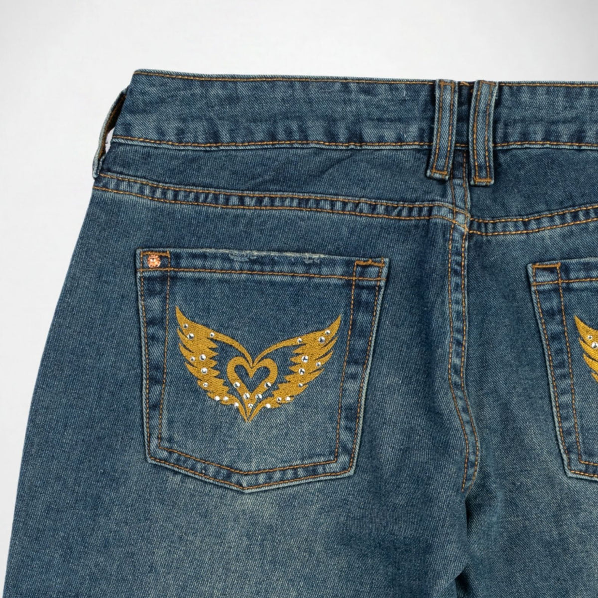 Close-up back view of blue denim Bubblegum jeans showing the back pockets with gold wing-and-heart embroidery and rhinestone detailing.