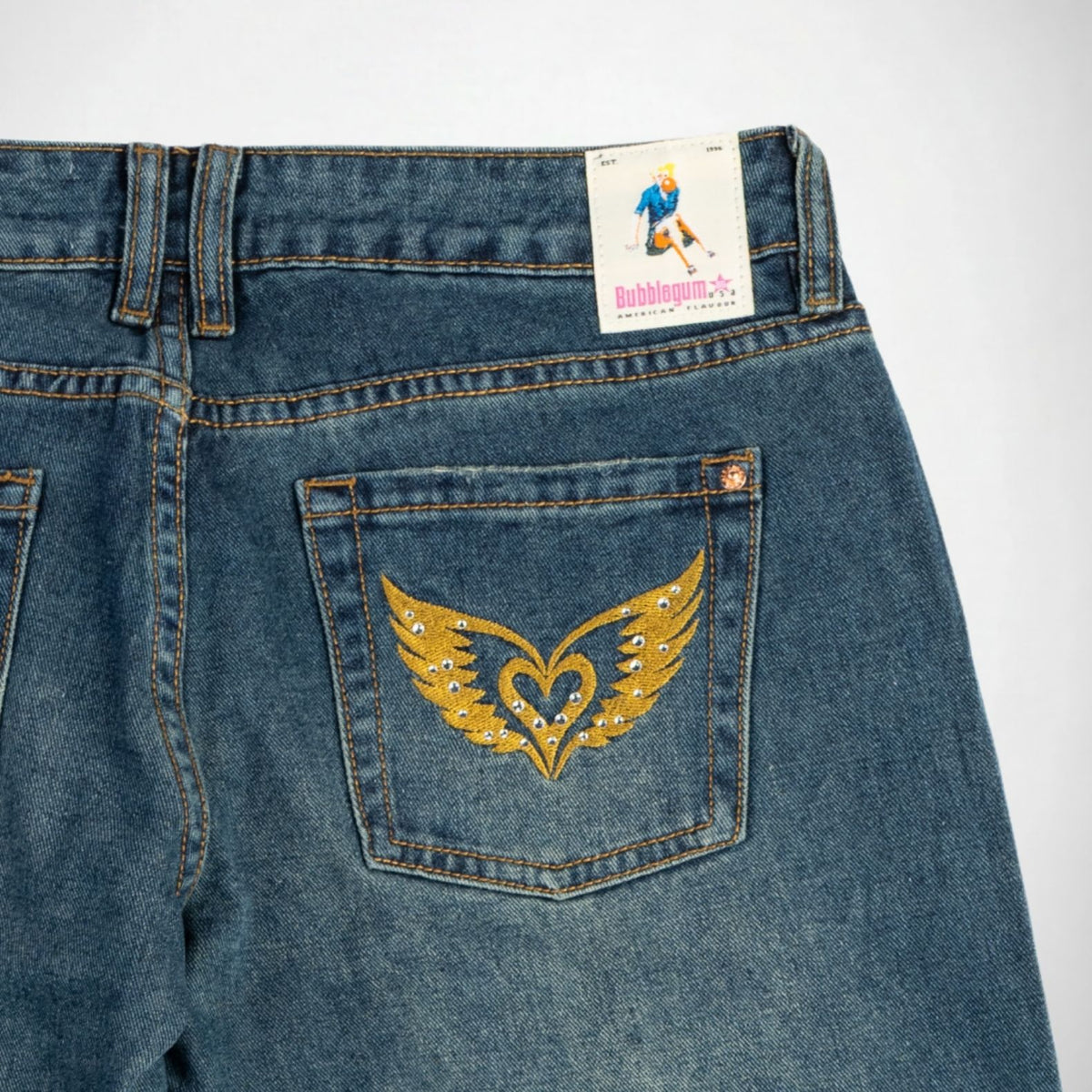 Close-up back view of blue denim Bubblegum jeans showing the back pocket with a gold embroidered winged heart design embellished with rhinestones, plus a visible Bubblegum USA brand label at the waistband.