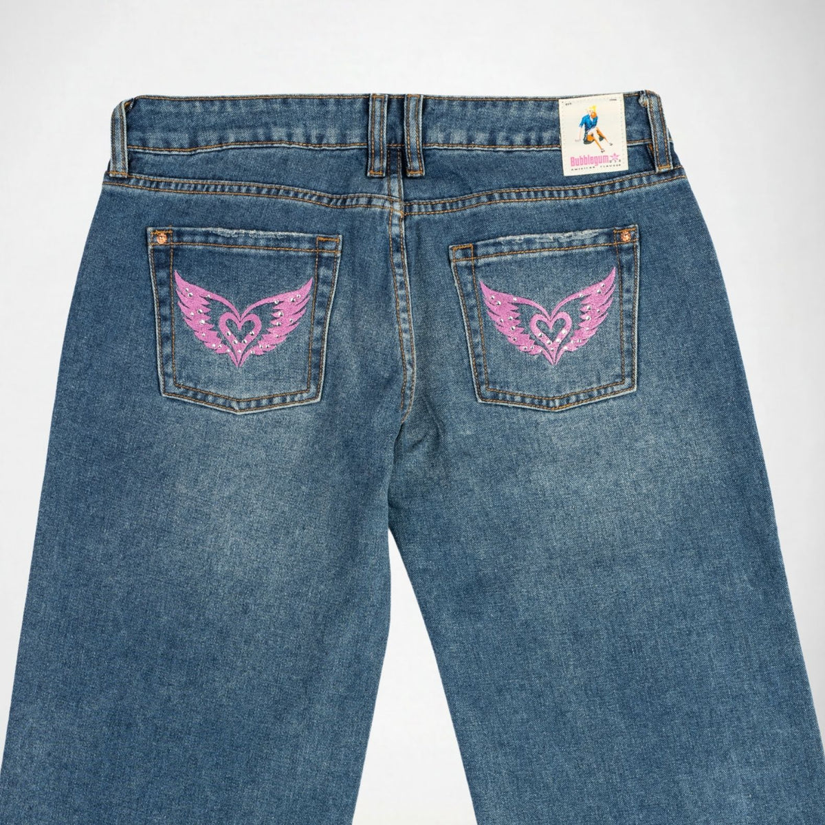 Back view of blue denim Bubblegum jeans featuring pink embroidered winged heart designs on both rear pockets, with a visible 'Bubblegum USA' label at the waistband.