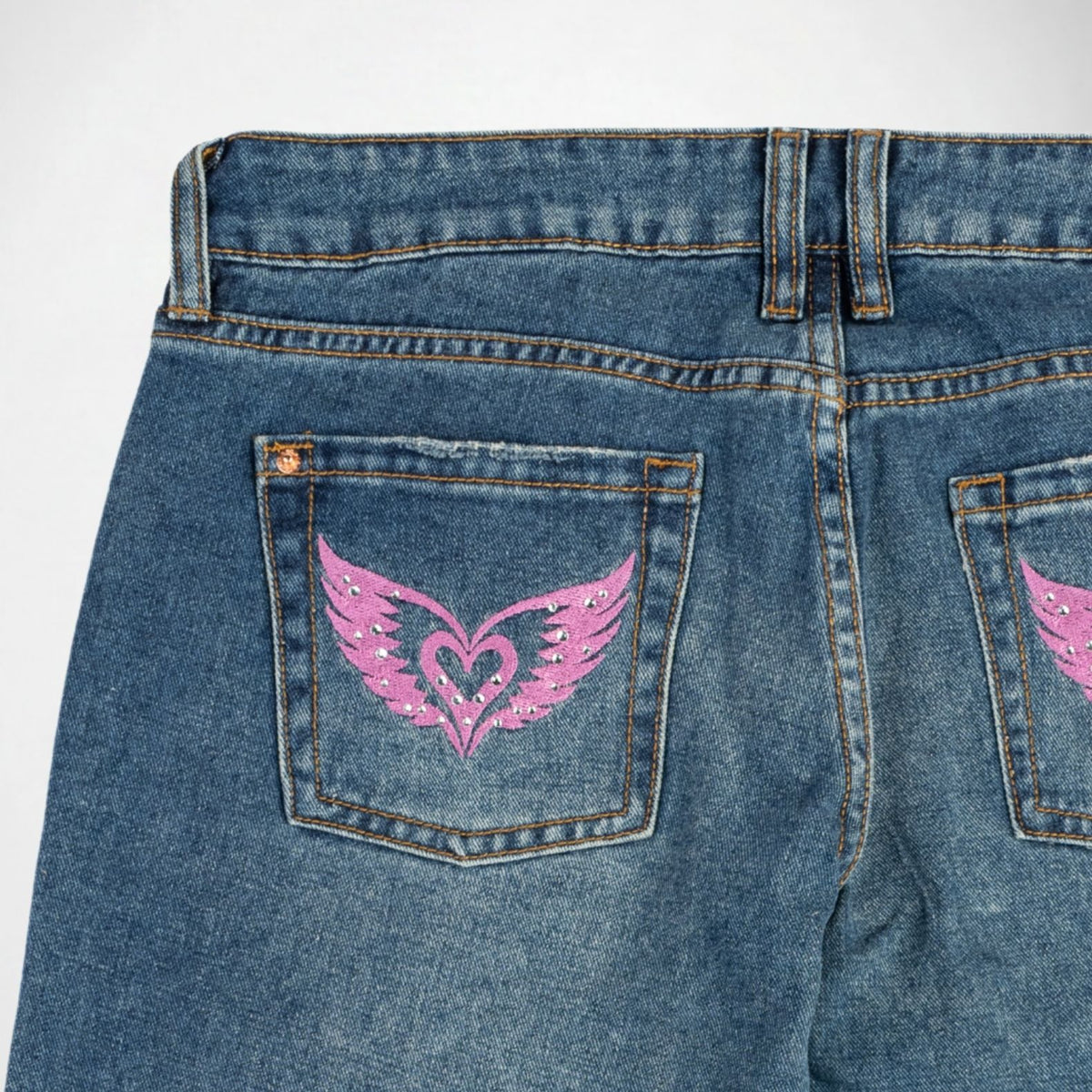 Close-up back view of blue denim Bubblegum jeans showing the back pockets with pink embroidered winged heart designs on a light gray background.