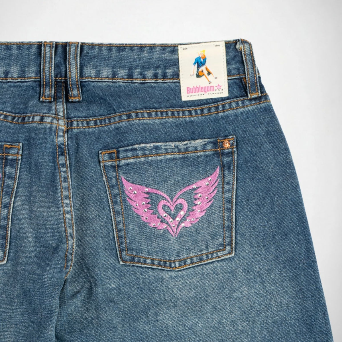 Close-up of the back of blue denim Bubblegum jeans showing a pink embroidered winged heart with rhinestones on the back pocket, with a visible Bubblegum USA brand label at the waistband.