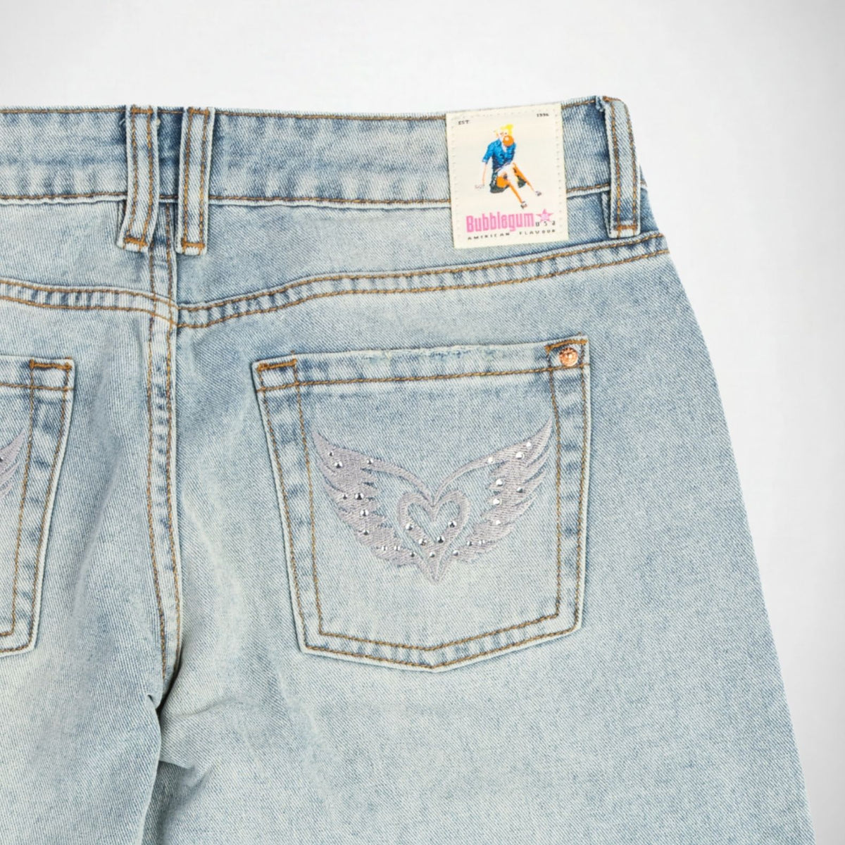 Close-up back view of light-wash blue denim Bubblegum jeans showing the waistband, belt loops, back pockets, and a silver wing-and-heart embroidery with rhinestones embellishment on the right pocket. A visible back label reads Bubblegum USA.