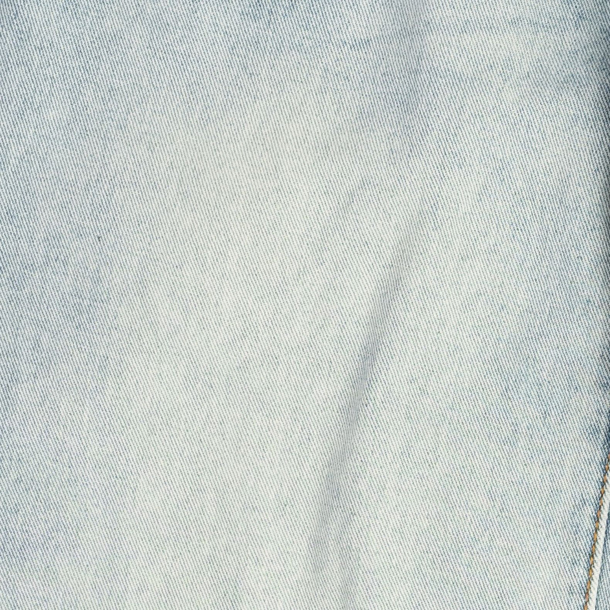 A close-up of light blue denim fabric texture with subtle fading and a diagonal seam visible on the right edge.