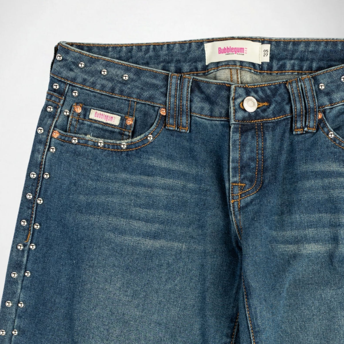 Close-up of a pair of blue denim Bubblegum jeans with silver studs embellishments along the pocket edges, orange contrast stitching, and a visible Bubbleum USA brand label.