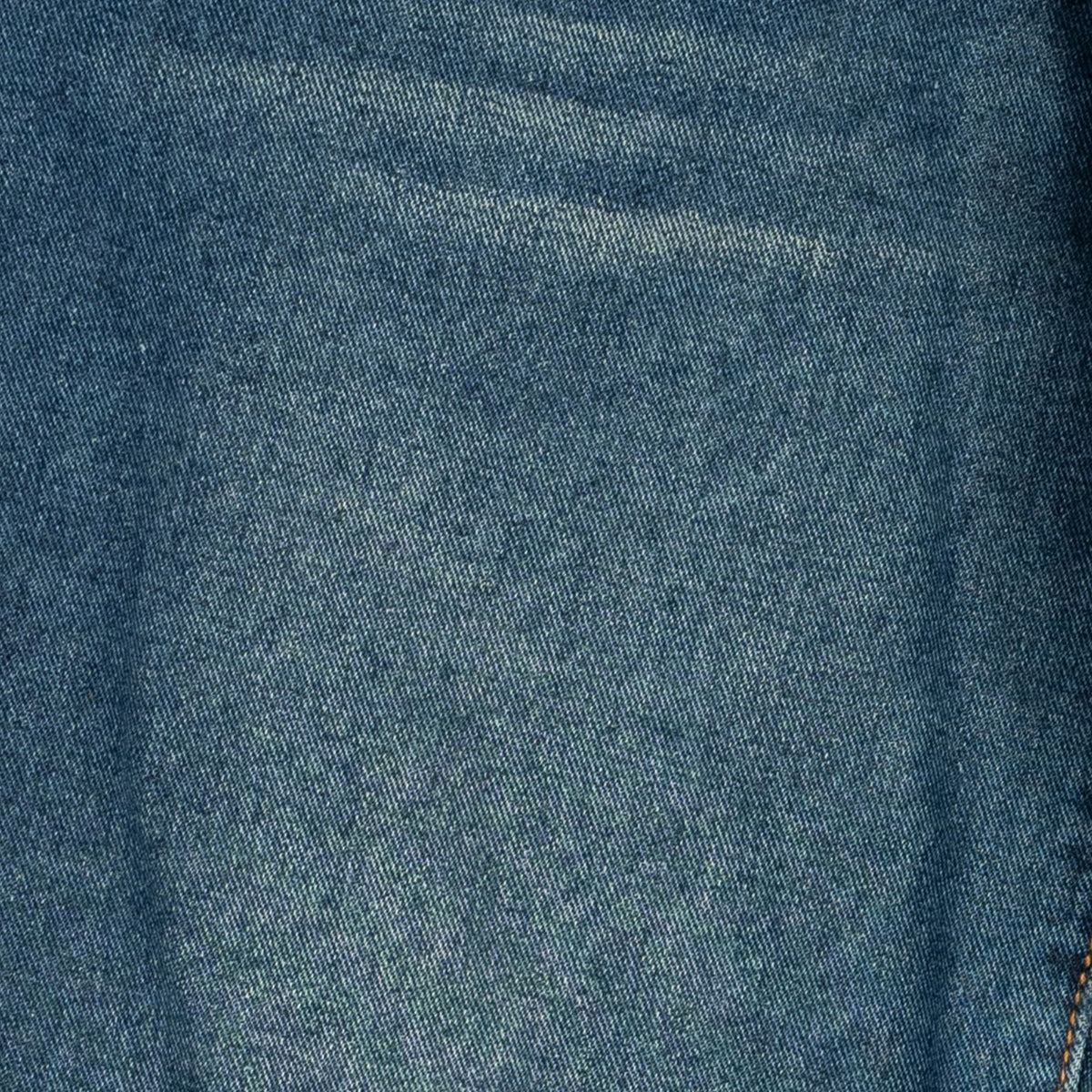 A close-up of blue denim fabric texture with subtle folds and a small portion of orange-brown stitching visible at the bottom right corner.